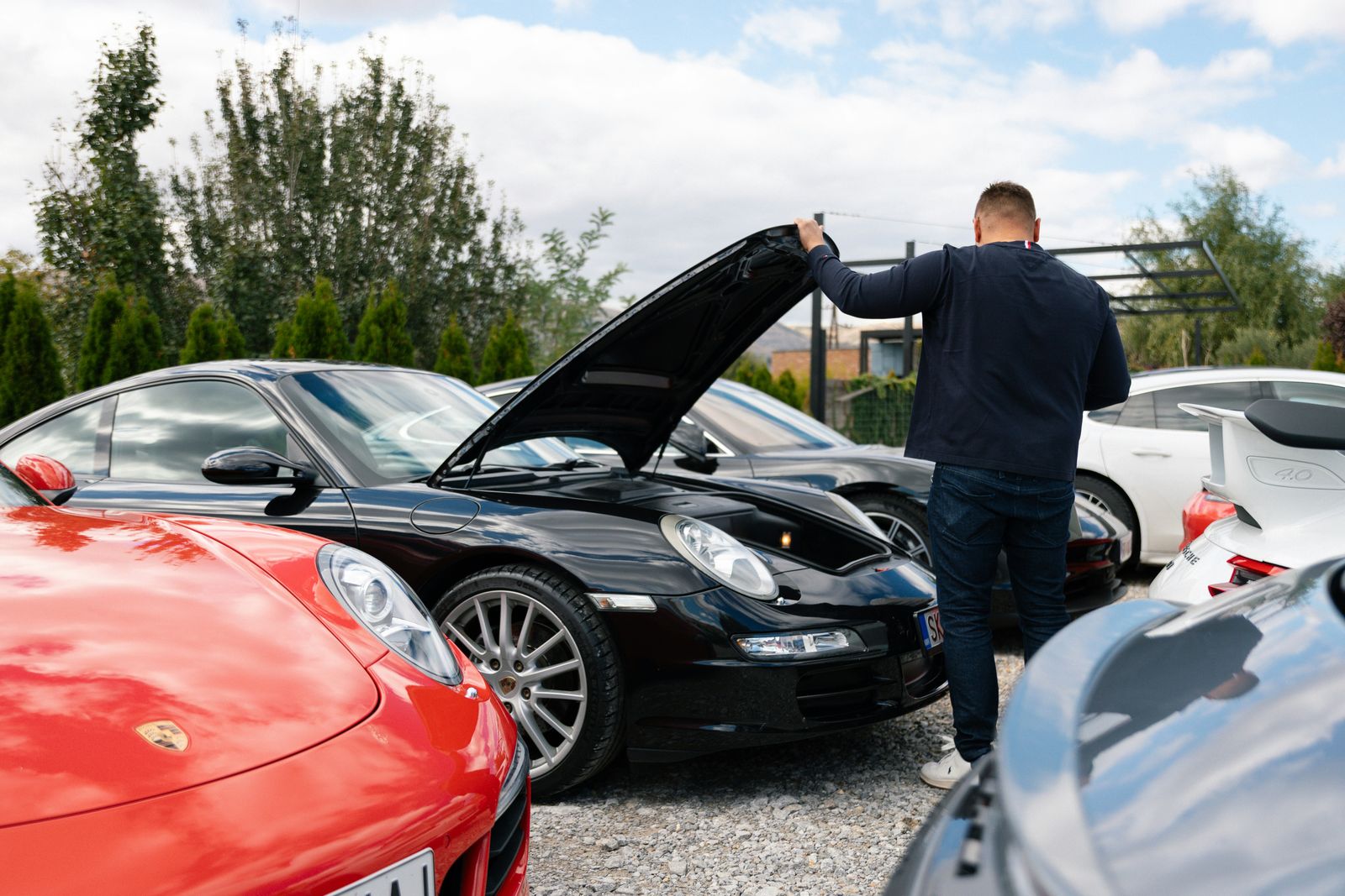 A man inspects a black sports car with its hood open in a lot filled with luxury cars.