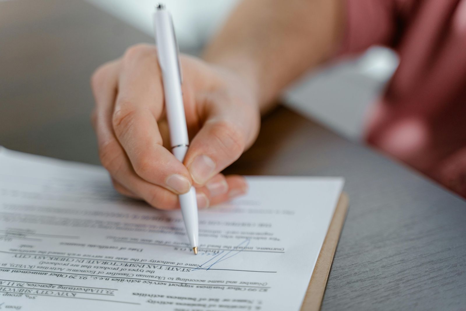 A close-up of a person’s hand holding a white pen and signing a document on a clipboard. The scene feels like a formal paperwork moment—finalizing terms, confirming consent, or approving a record.