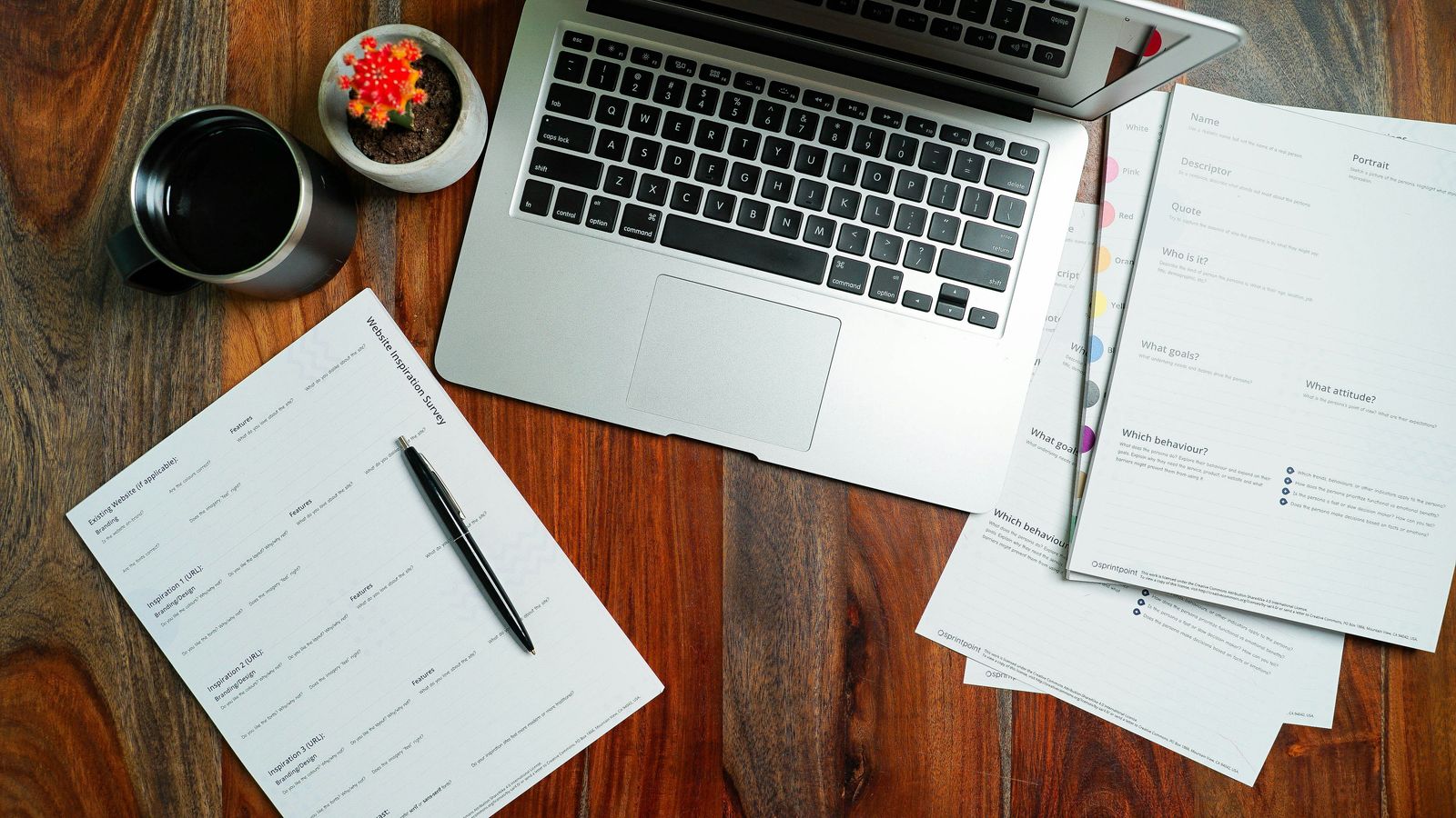 Overhead view of a laptop, printed planning worksheets, pen, coffee mug, and small plant on a wooden desk.