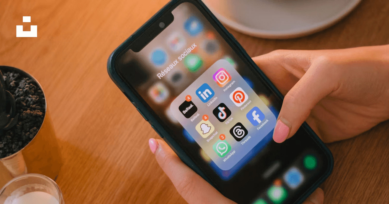 A close-up of someone holding a smartphone with a “Social Networks” folder open, showing apps like Instagram, LinkedIn, TikTok, Facebook, and WhatsApp.