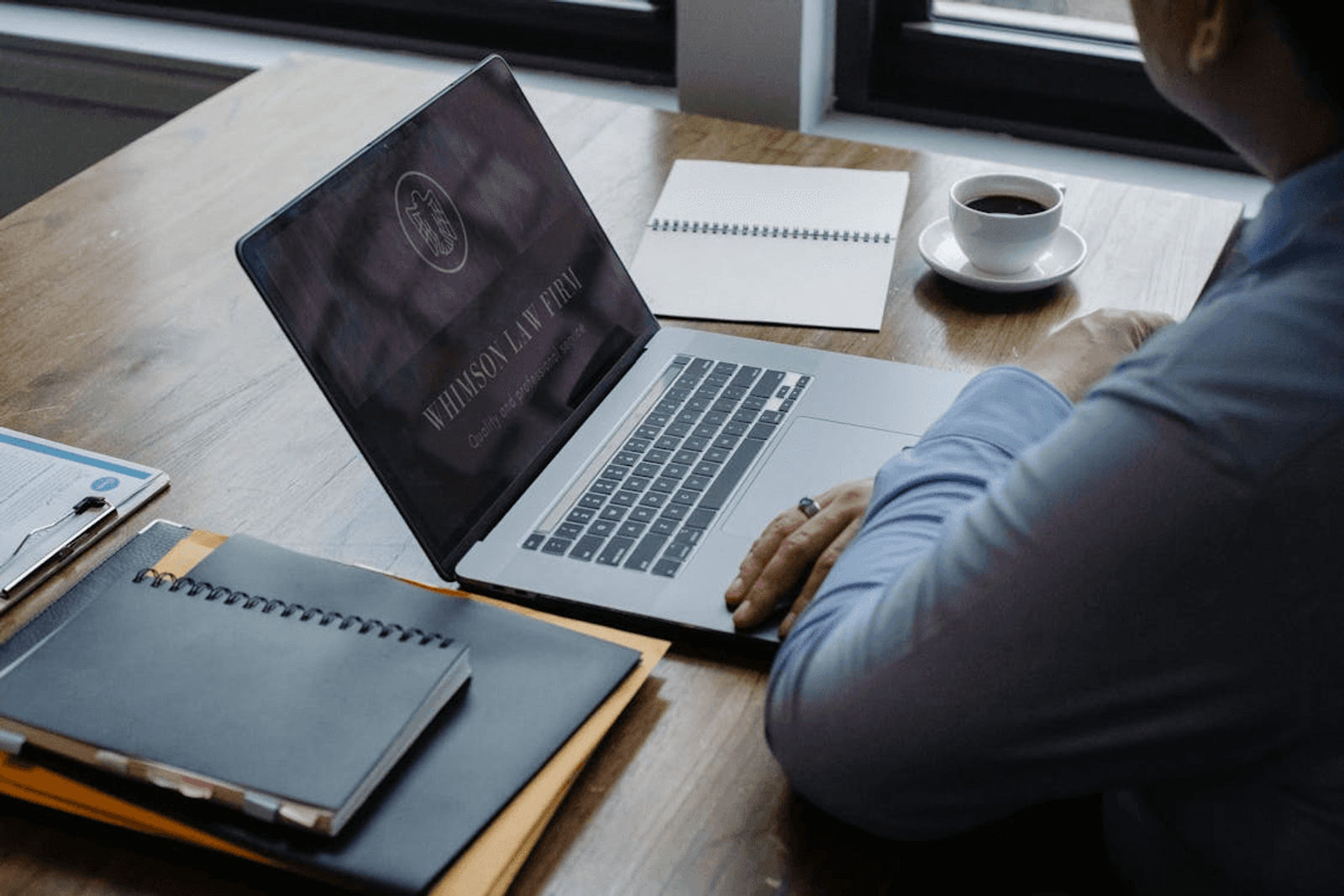 A lawyer-style workstation scene: laptop open with a law firm screen, paperwork and a clipboard on the side, and coffee on the table by a window.