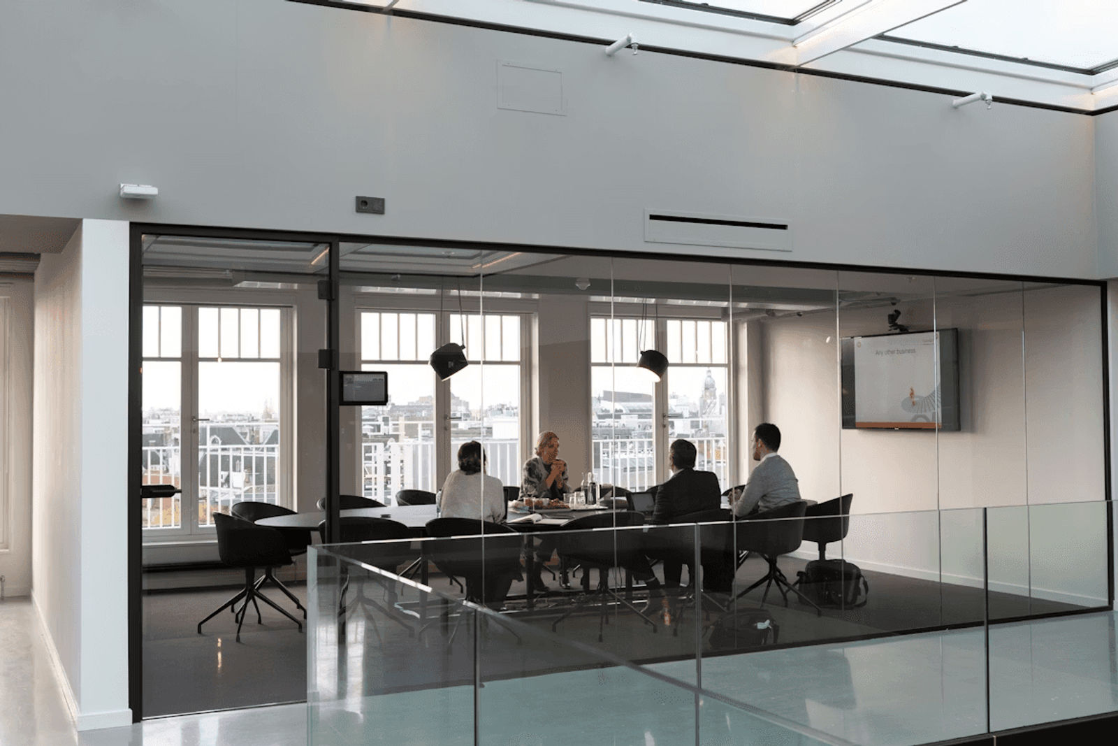 A small group meeting in a glass-walled conference room, with people seated around a table and a presentation screen on the wall.