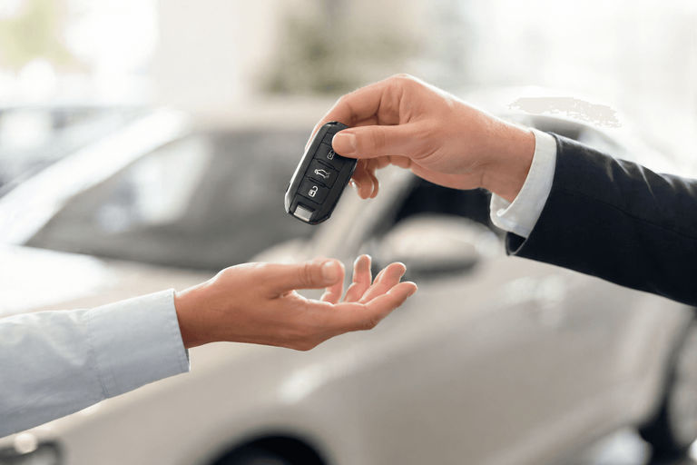 Close-up of one person handing a car key fob to another in a bright car showroom, with a blurred vehicle in the background.
