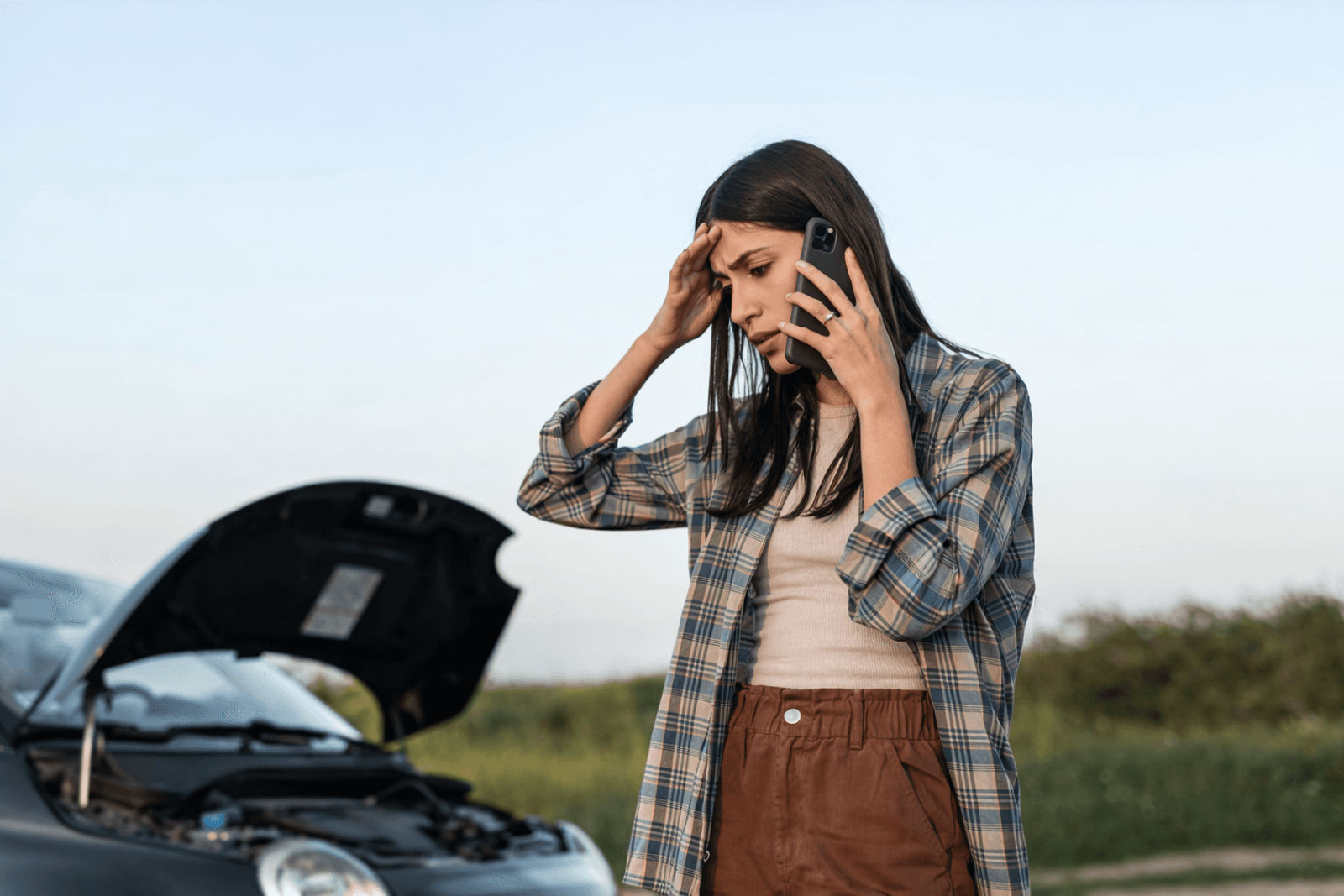 A worried woman on the phone stands beside a car with the hood up on a roadside, looking stressed as if she’s dealing with a breakdown.