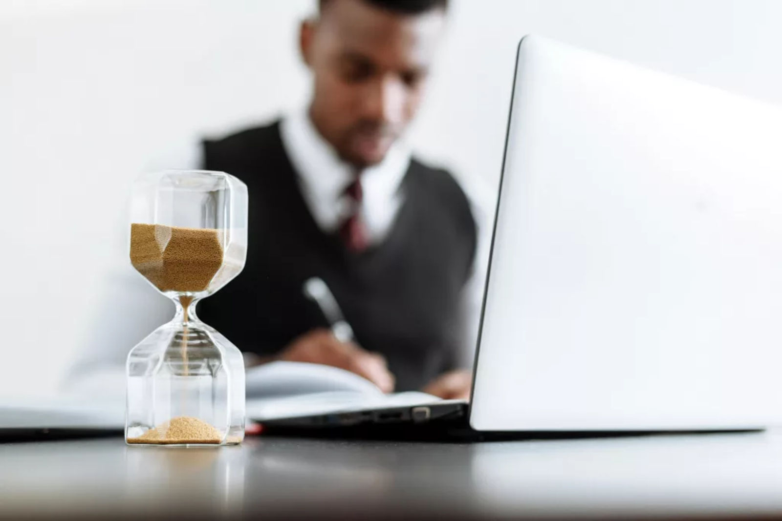 Hourglass on a desk with a person working on a laptop in the background