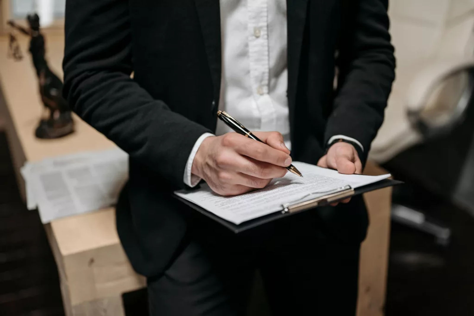 Person in formal attire writing on a clipboard near legal documents