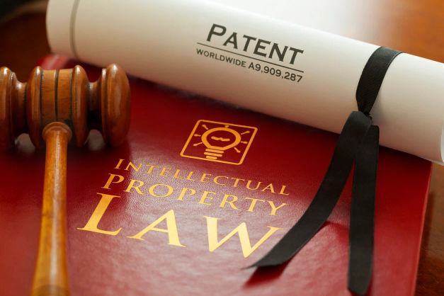A close-up of a rolled PATENT certificate tied with a ribbon, resting on a red “Intellectual Property Law” book, with a judge’s gavel beside it. It’s a classic visual for protecting inventions and enforcing ownership rights through the legal system.