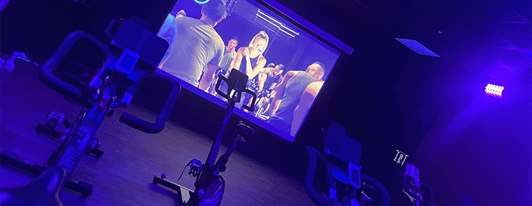 indoor cycling bikes in a lowly lit room with a screen at the front with virtual instructor