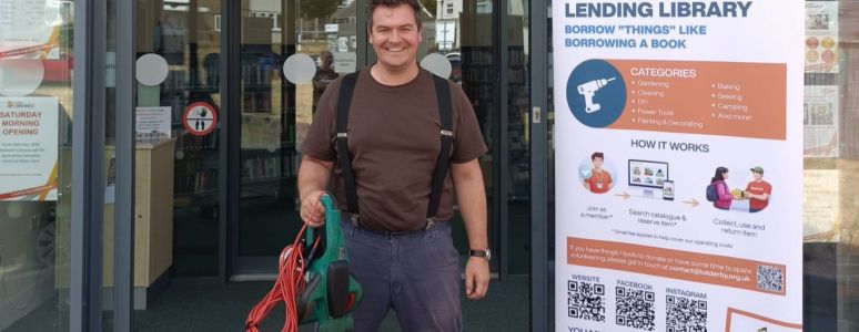 A man stands in front of Chellaston Library with gardening tools, next to a Library of Things pull-up banner