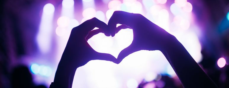 Silhouette of two hands in the air making a heart shape with bright lights from stage in the background
