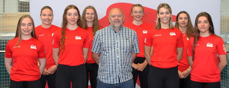 Cllr Jerry Pearce surrounded by women from Team England in red cycling jerseys against a red and white backdrop