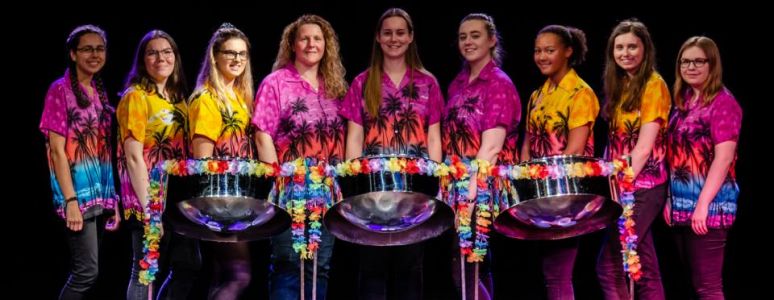 Members of Pure Steel wearing yellow and pink shirts, standing behind a line of steel drums