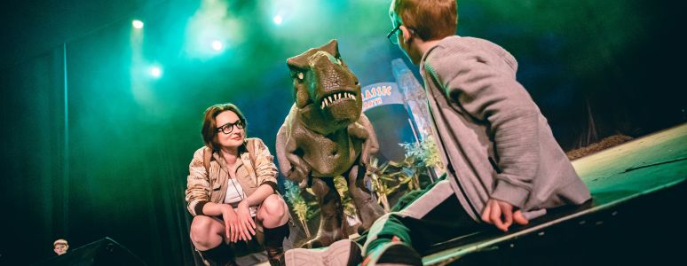 An animatronic dinosaur stands beside a ranger and looks at a child on stage.