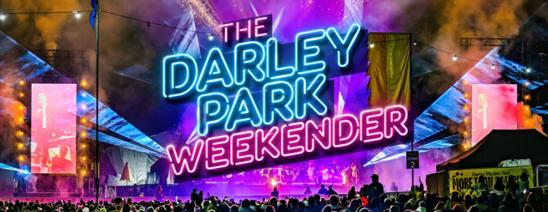 A night-time view of The Darley Park Weekender festival with a large neon sign, fireworks, and a crowd facing the stage.