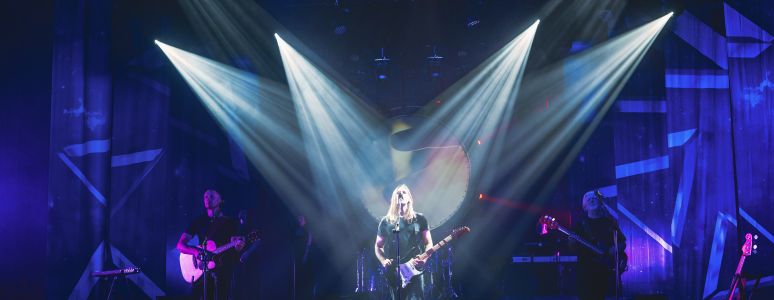 Three guitarists stand on stage. The lead is highlighted by many spotlights as he sings into the microphone.