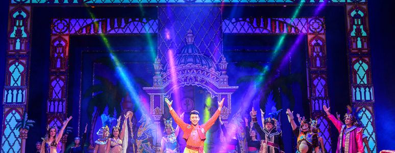 Aladdin stands on stage with his arms in the air.