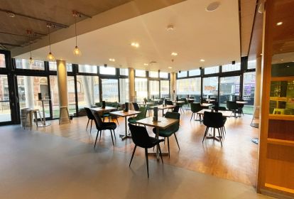 Lounge var area set with several small tables with green padded chairs.  Two walls are made of glass so the room looks out into Derby market place.