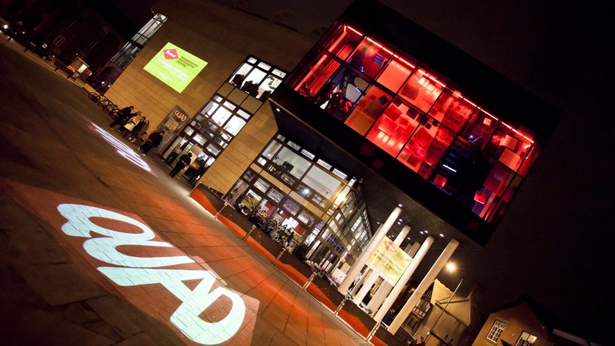 The exterior of the QUAD building at night with QUAD lit up my red lights and the QUAD logo projected onto the ground in front of the building