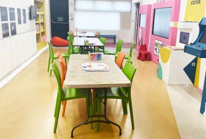 A brightly coloured room with activity tables set down the centre and interactive screens on the walls