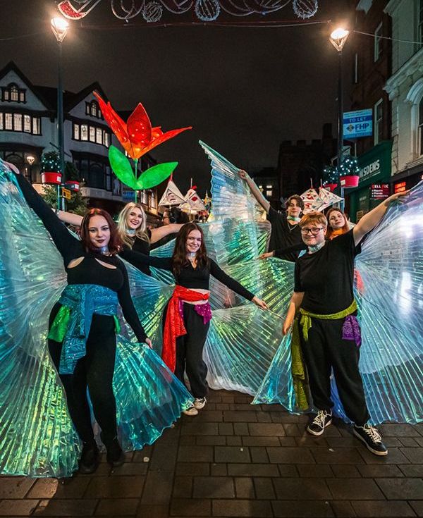 A lively group of people in colorful costumes, each holding vibrant wings, smiling and posing together.