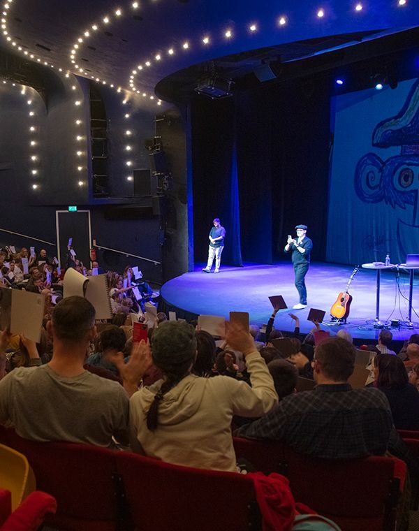 A lively crowd in a theater, captivated by a concert performance on stage, with colorful lights illuminating the scene.
