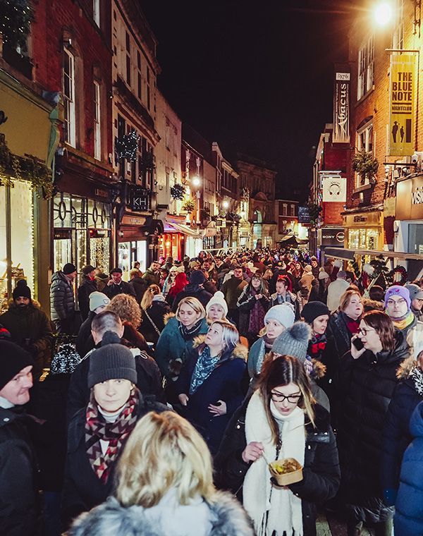 A crowd of warmly dressed people fill Sadler Gate at night