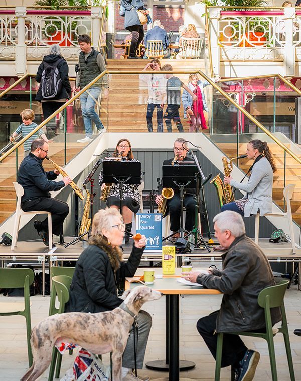 A lively scene at Derby Market Hall of people at tables enjoying time with dogs while musicians play nearby, creating a vibrant atmosphere.