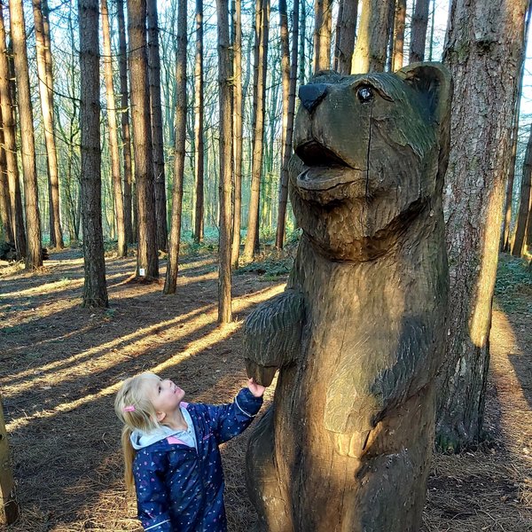 A little girl standing next to a wooden bear statue in a woodland