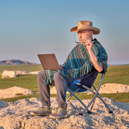 An older man works on a laptop outdoors in a remote rural landscape at sunset