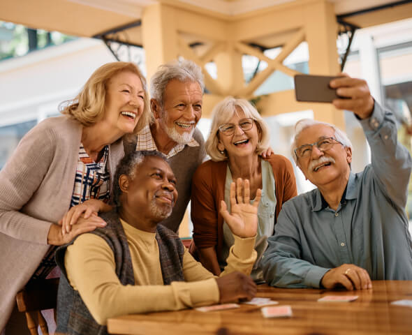Group of five smiling older friends taking a selfie together at a card table