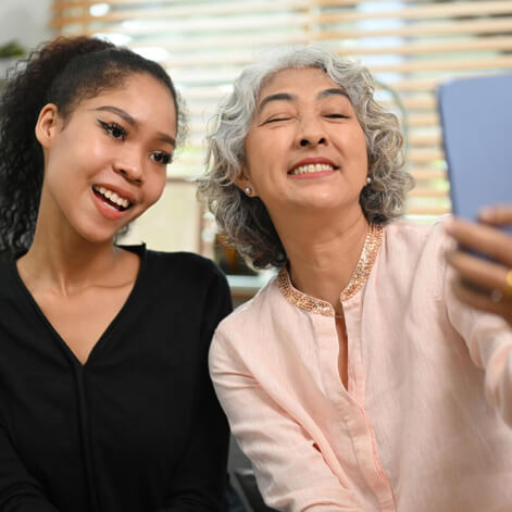 A senior woman and her granddaughter smile together at a phone screen at home.