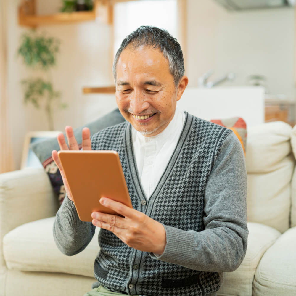 Smiling older man in a gray cardigan waving at a tablet during a video call at home