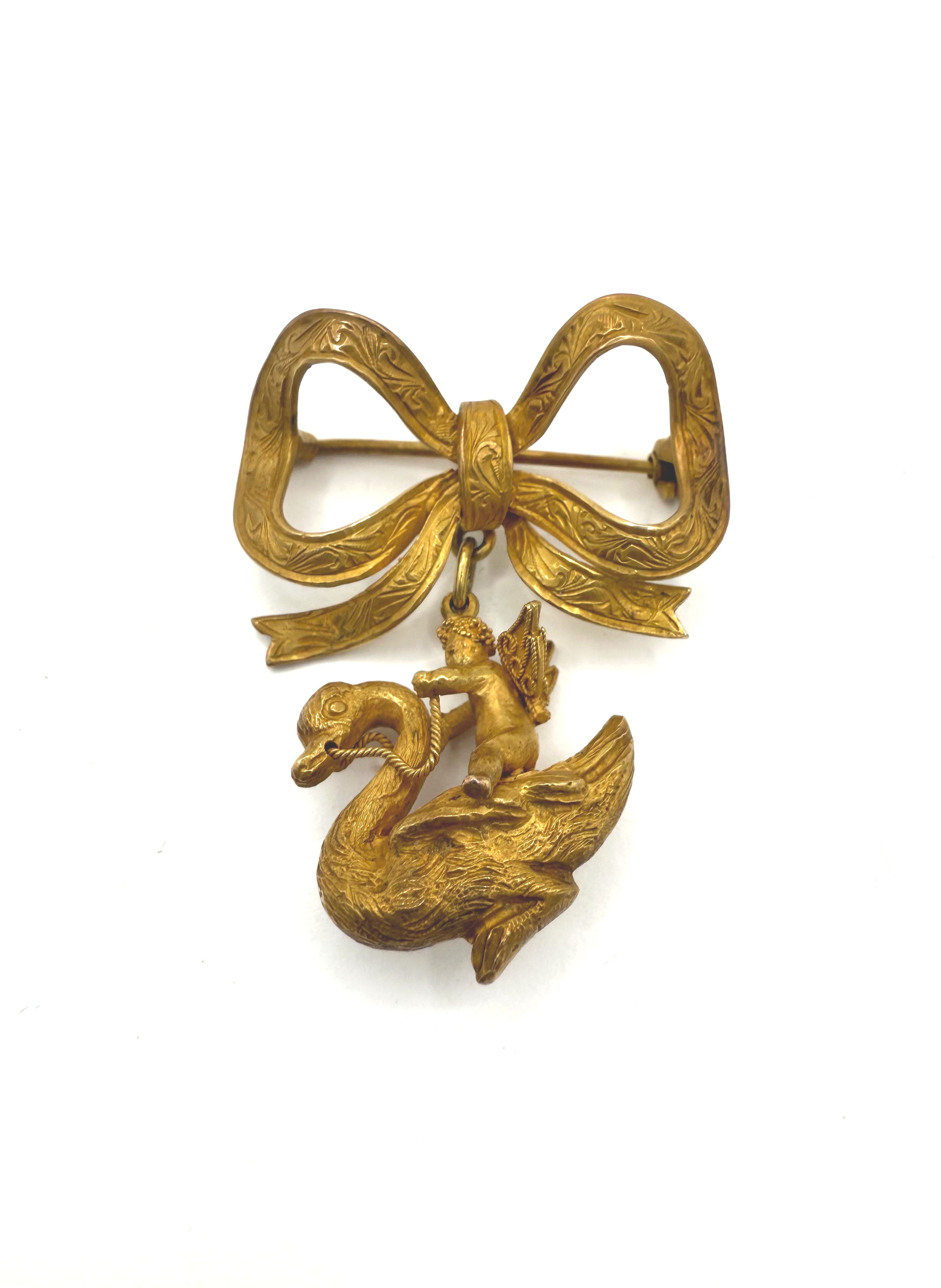 Broche en or jaune (750 et 585 millièmes) amati stylisant un noeud et retenant un angelot sur un cygne. - Lazuli