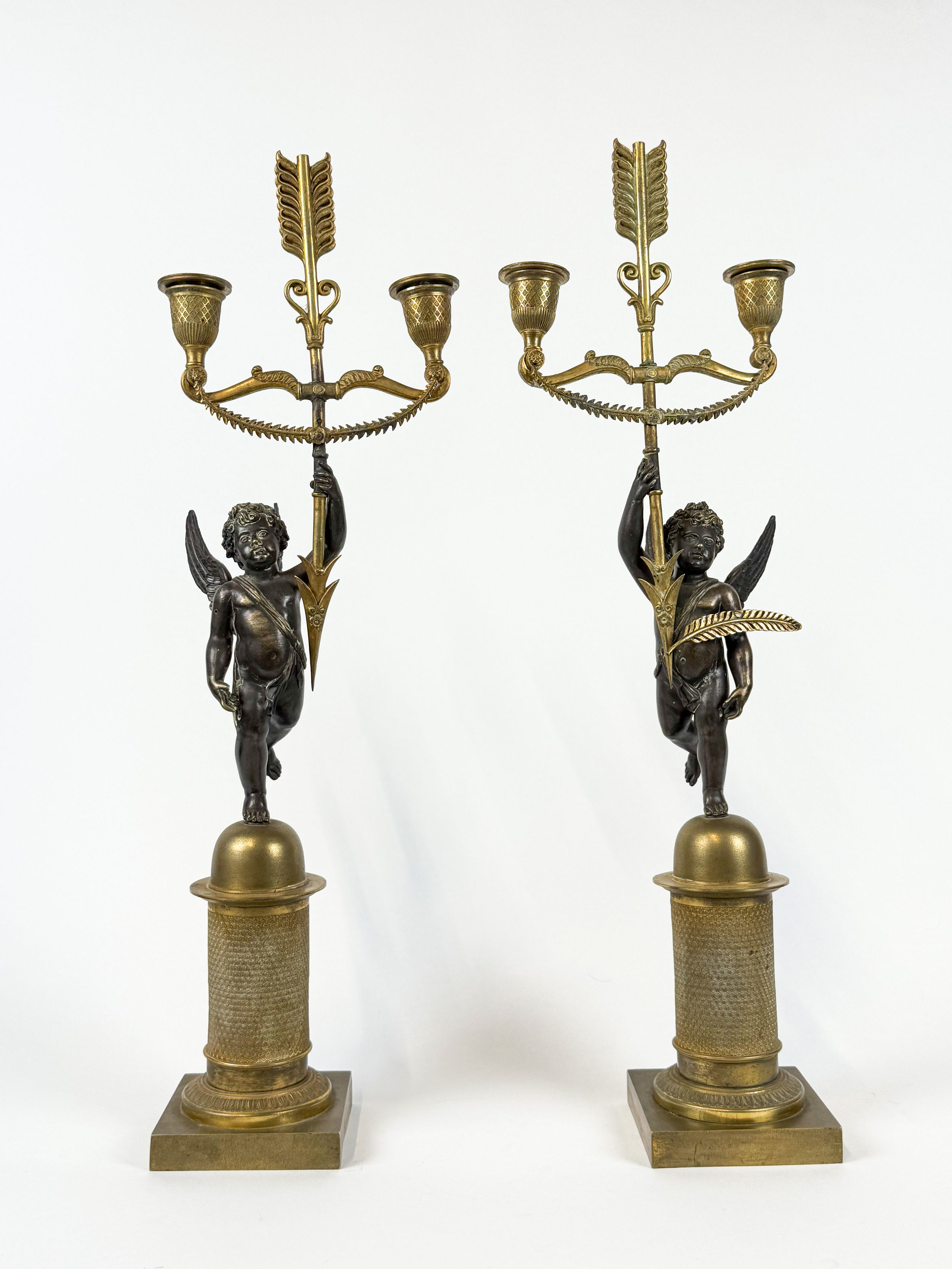 Paire de candélabres en bronze à patine noire et dorée à deux feux de lumière représentant des angelots retenant une flèche porte-torchères. - Lazuli