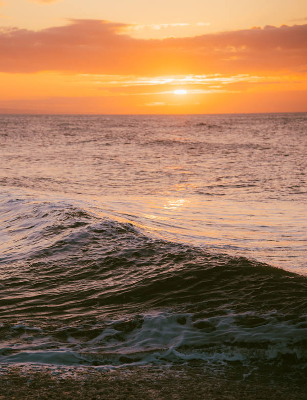 A wave curling toward the shore at sunset, the sun a bright disc on the horizon and the sea lit gold and amber