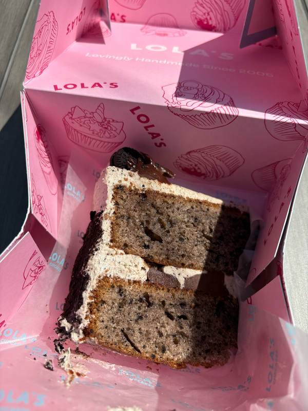 A thick slice of cookies-and-cream layer cake propped inside a bright pink Lola's Cupcakes box, shot in hard sunlight