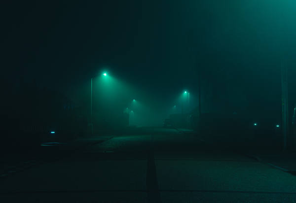 A village street at night in thick fog, two streetlights burning green haloes in the mist and a figure barely visible between them