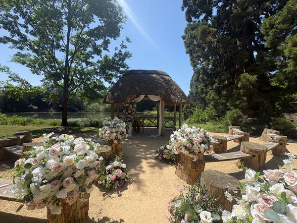 An outdoor wedding ceremony set up in a garden: a thatched wooden pavilion at the end of a gravel aisle, flanked by benches and tall pale flower arrangements