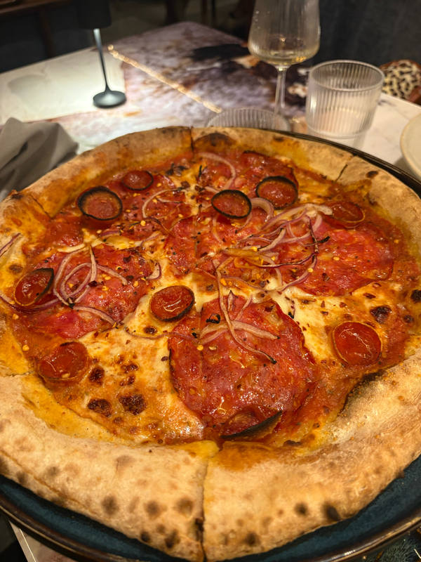 A whole pepperoni pizza on a dark metal pan in a restaurant, blistered crust and glasses on the table behind