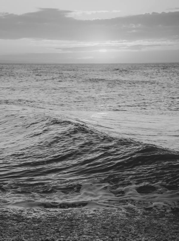 A black and white photograph of a single wave rising over a shingle beach, the sun a pale smudge behind cloud