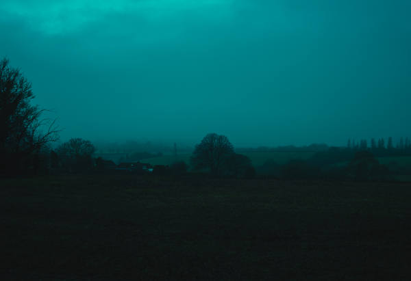 A dark field at dusk under heavy cloud, the whole frame washed teal, a low treeline and distant lights on the horizon