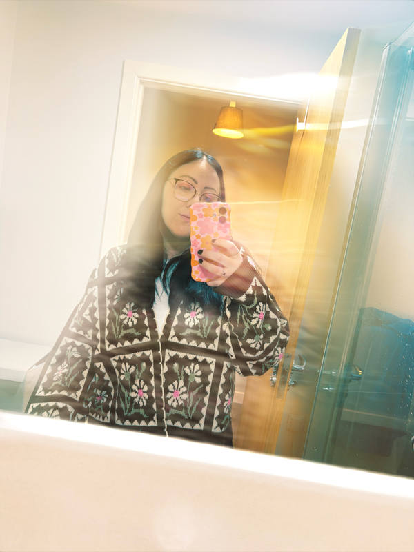 A mirror selfie taken through a doorway: a woman in a black and white patterned jacket holding up a pink phone, warm lamplight behind her