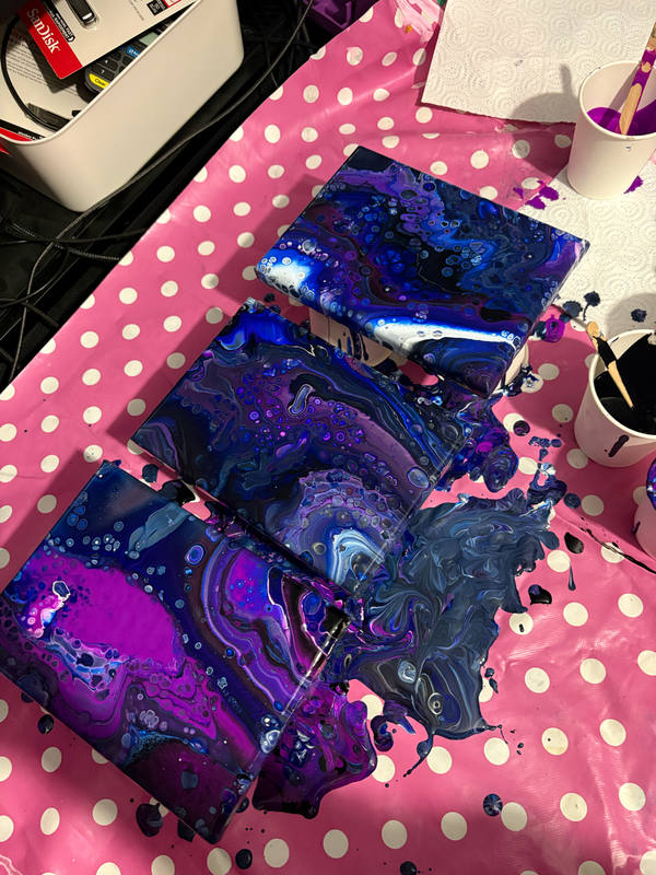 Four small finished pour paintings in violet, cobalt and black laid out to dry on a pink polka-dot sheet, paint pots and brushes around them