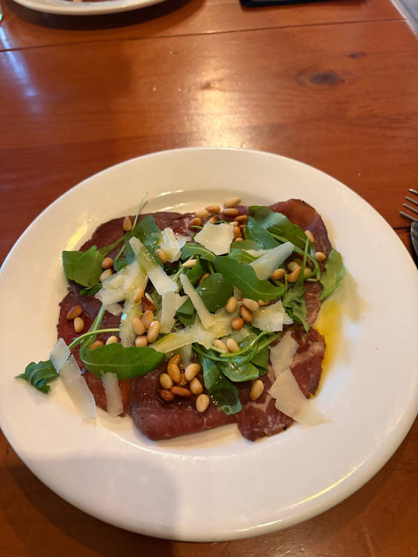 A plate of beef carpaccio topped with rocket, shaved parmesan and pine nuts, on a wooden restaurant table