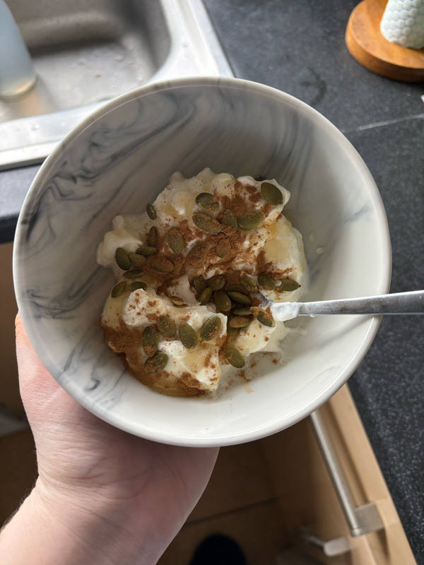 A marbled ceramic bowl held over a kitchen worktop, filled with plain yoghurt, pumpkin seeds and a dusting of cinnamon, spoon resting inside