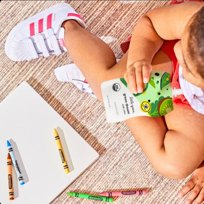 Baby sitting with crayons and a notebook while drinking a Little Spoon Smoothie pouch