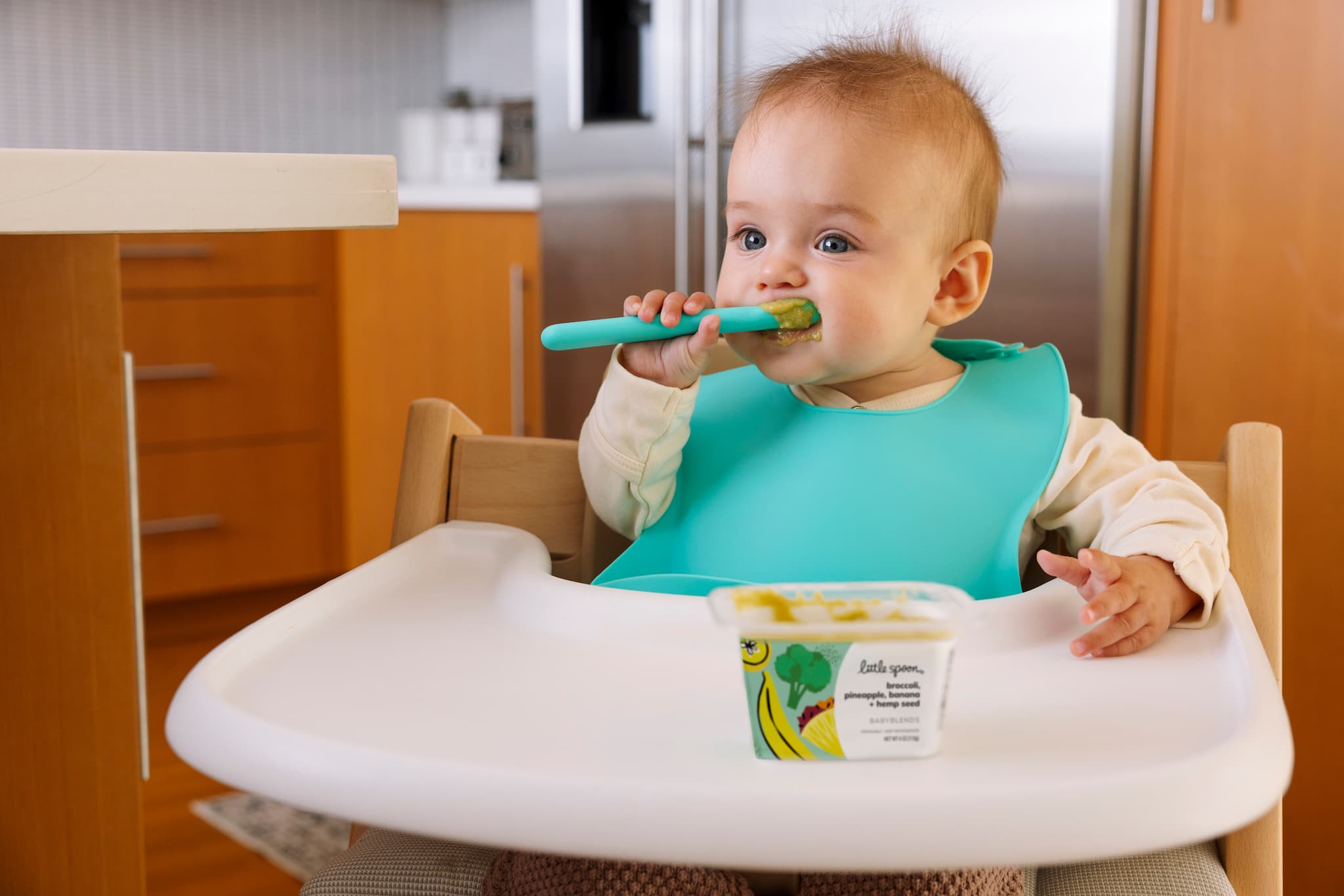 Baby eating from a spoon while sitting in a highchair with a Little Spoon Babyblends cup on the tray.