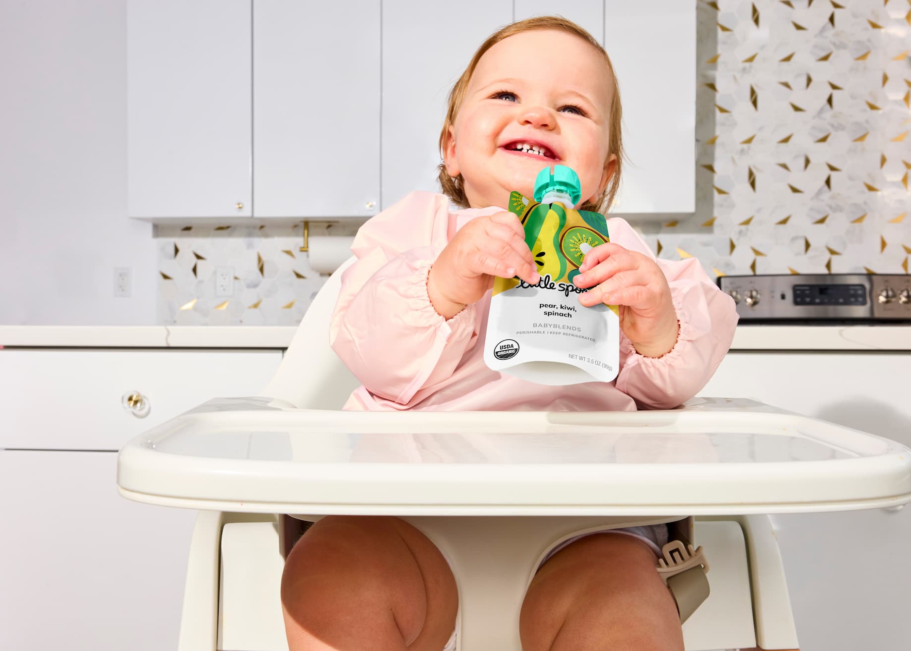 Smiling baby holding and eating from a Little Spoon Babyblends pouch in a highchair.
