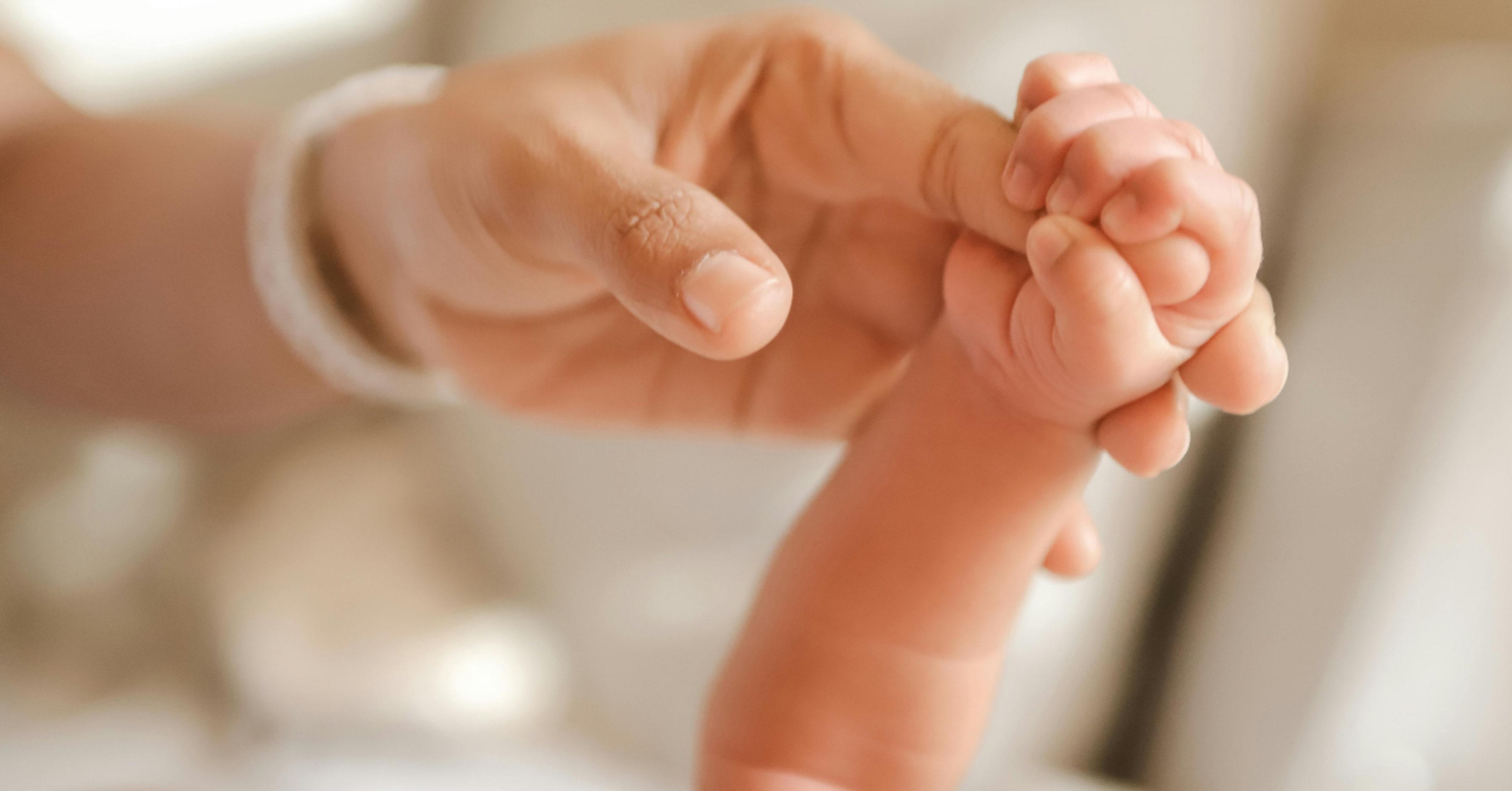Close-up of a newborn baby gently holding an adult’s finger, with a soft, neutral background.
