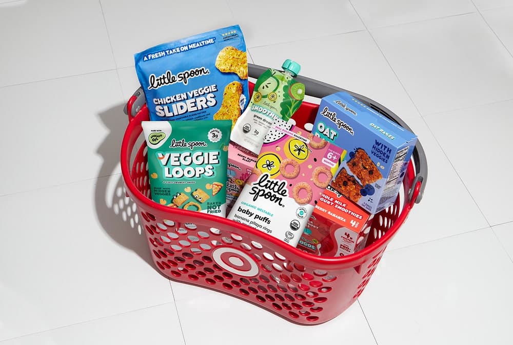 Red Target shopping basket filled with various Little Spoon kids’ food products, including Veggie Loops, Baby Puffs, Smoothies, Chicken Veggie Sliders, and Oat Bakes, placed on a white tiled floor